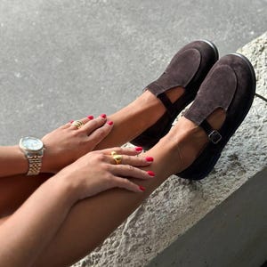 May include: Dark brown suede Mary Jane style shoes with a strap and buckle closure. The shoes have a rounded toe and a low heel. A gold watch and rings are also visible.
