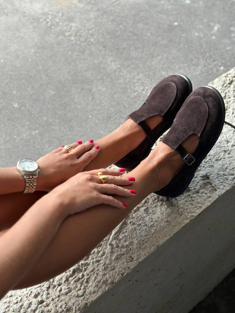 May include: Dark brown suede Mary Jane style shoes with a strap and buckle closure. The shoes have a rounded toe and a low heel. A gold watch and rings are also visible.