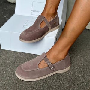 May include: Taupe suede flats with a T-strap design and a buckle closure. The shoes have a rounded toe and a light-colored sole. The brand name "Maletskiy" is visible on the box in the background. These flats are a stylish footwear option.