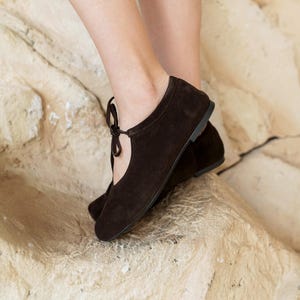 May include: A pair of dark brown suede ballet flats with a rounded toe and a delicate tie detail. The shoes have a low heel and a simple, elegant design, perfect for everyday wear. The flats are shown on a textured, light-colored surface.