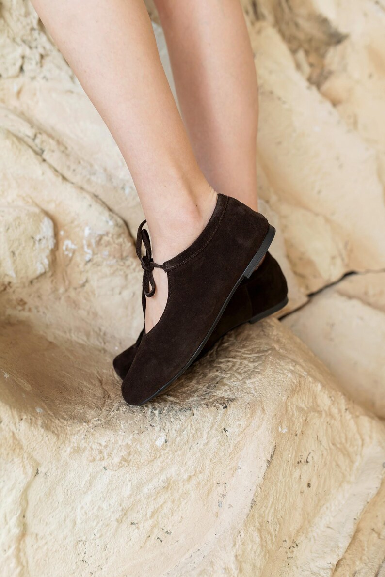 May include: A pair of dark brown suede ballet flats with a rounded toe and a delicate tie detail. The shoes have a low heel and a simple, elegant design, perfect for everyday wear. The flats are shown on a textured, light-colored surface.