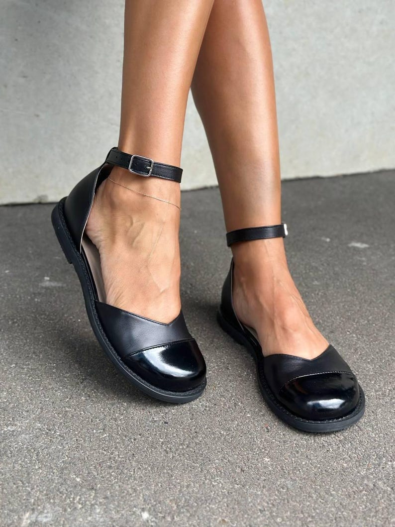 Puede incluir: Un par de zapatos planos negros con punta redondeada de acabado brillante, parte superior de cuero mate y una fina correa al tobillo con hebilla de metal plateado. Los zapatos tienen un dise&ntilde;o de perfil bajo con una suela negra s&oacute;lida.