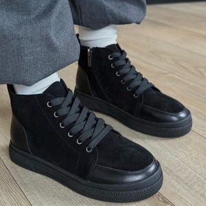 Könnte beinhalten: Ein Paar schwarze High-Top-Sneaker aus einer Kombination von Wildleder- und Glattledereinsätzen. Die Schuhe verfügen über schwarze Schnürsenkel, silberfarbene Ösen, einen seitlichen Reißverschluss und eine strukturierte schwarze Gummisohle.