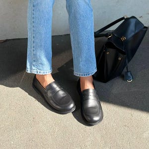 Puede incluir: Un par de mocasines de cuero negro con un diseño clásico. Los zapatos se usan con vaqueros azules claros. También se ve un bolso negro con un cierre dorado.