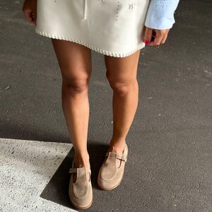 May include: A pair of light brown suede Mary Jane shoes with a buckle closure. The shoes have a rounded toe and a small heel. The image also shows a white skirt with decorative stitching and a light blue long-sleeve shirt.