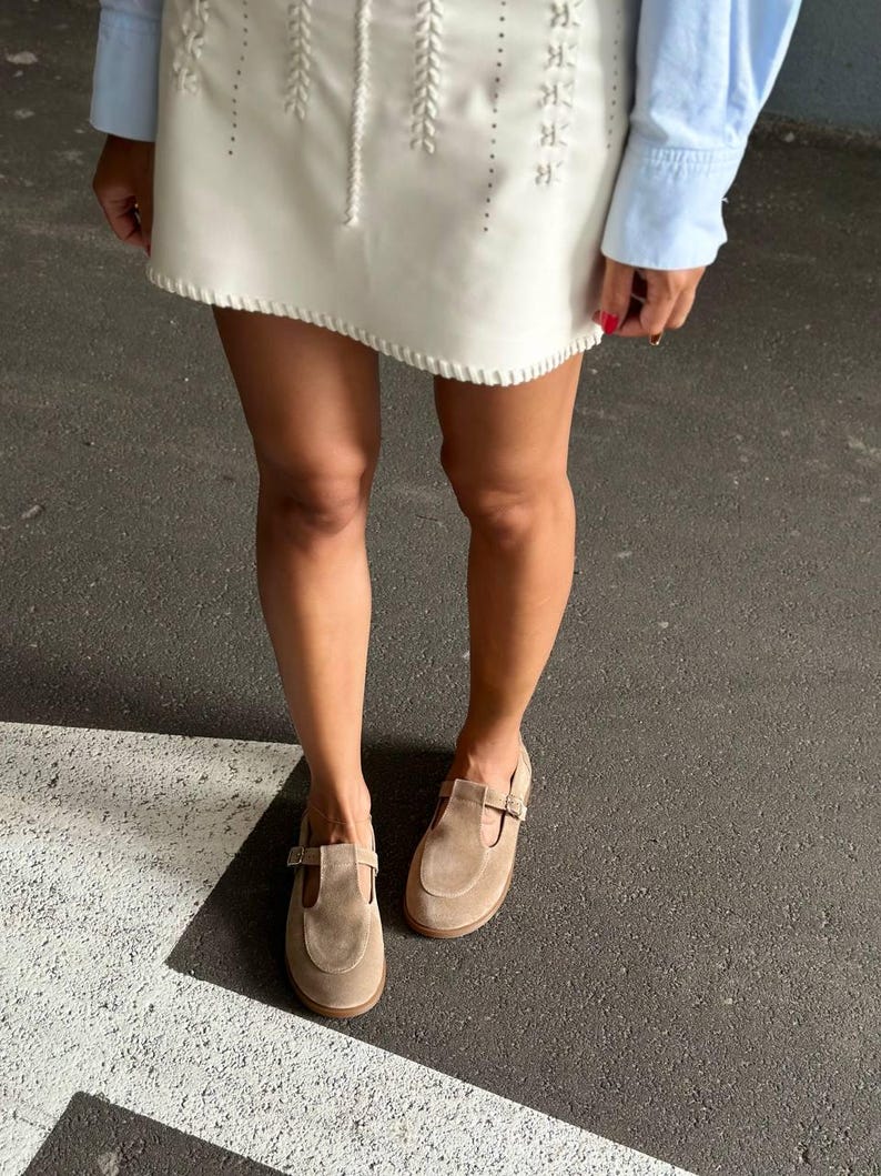 May include: A pair of light brown suede Mary Jane shoes with a buckle closure. The shoes have a rounded toe and a small heel. The image also shows a white skirt with decorative stitching and a light blue long-sleeve shirt.