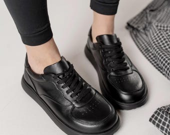 May include: A pair of black leather sneakers with black laces. The shoes have a thick, black sole and are worn with black pants. A black and white plaid pattern is visible in the background.