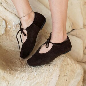 May include: A pair of dark brown suede ballet flats with a tie closure. The shoes have a rounded toe and a low heel. The image shows the shoes on a light-colored, textured surface.