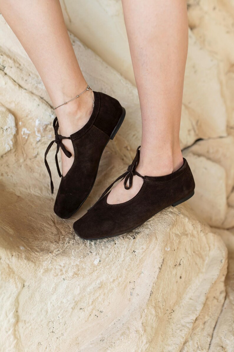 May include: A pair of dark brown suede ballet flats with a tie closure. The shoes have a rounded toe and a low heel. The image shows the shoes on a light-colored, textured surface.