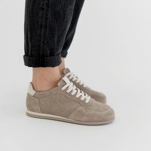 Pu&ograve; includere: Sneakers in camoscio color talpa con lacci bianchi e un inserto sul tallone color crema. Le scarpe basse hanno un design classico con suola in gomma. Le sneakers sono mostrate con jeans in denim grigio scuro.