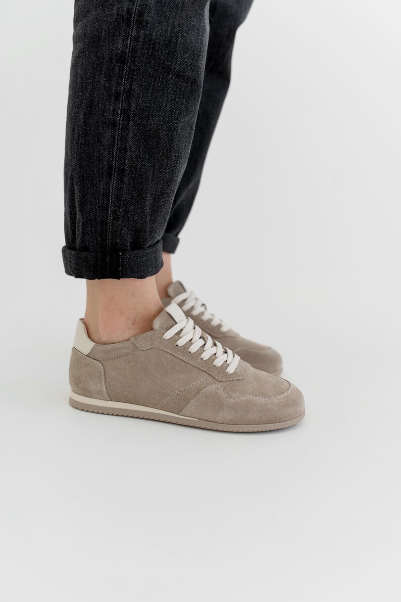 Pu&ograve; includere: Sneakers in camoscio color talpa con lacci bianchi e un inserto sul tallone color crema. Le scarpe basse hanno un design classico con suola in gomma. Le sneakers sono mostrate con jeans in denim grigio scuro.