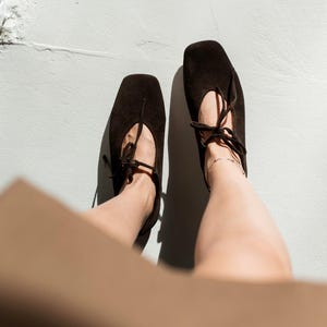 May include: A pair of dark brown suede ballet flats with square toes and delicate tie closures. The shoes are positioned on a light gray surface, with shadows cast by the sunlight. The overall aesthetic is minimalist and chic.