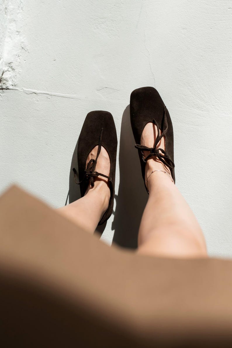 May include: A pair of dark brown suede ballet flats with square toes and delicate tie closures. The shoes are positioned on a light gray surface, with shadows cast by the sunlight. The overall aesthetic is minimalist and chic.