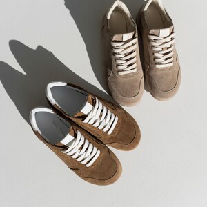 Pu&ograve; includere: Due paia di sneakers in pelle scamosciata marrone con lacci bianchi e dettagli bianchi. Le sneakers sono esposte su una superficie bianca, con ombre dalla luce solare. Scarpe dallo stile casual.