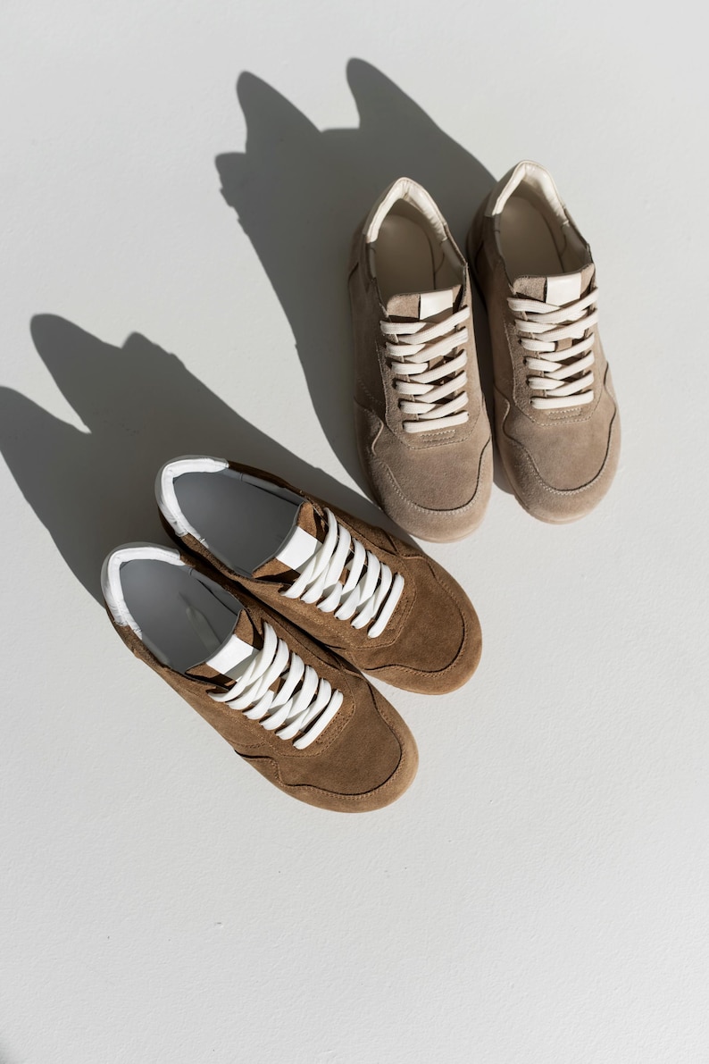 Pu&ograve; includere: Due paia di sneakers in pelle scamosciata marrone con lacci bianchi e dettagli bianchi. Le sneakers sono esposte su una superficie bianca, con ombre dalla luce solare. Scarpe dallo stile casual.
