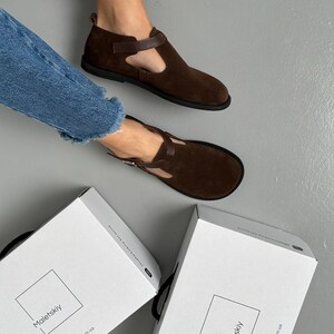 May include: Brown suede T-strap shoes with a dark brown strap and buckle closure. The shoes have a rounded toe and a black sole. Two white boxes with the brand name "Maletskiy" are visible.