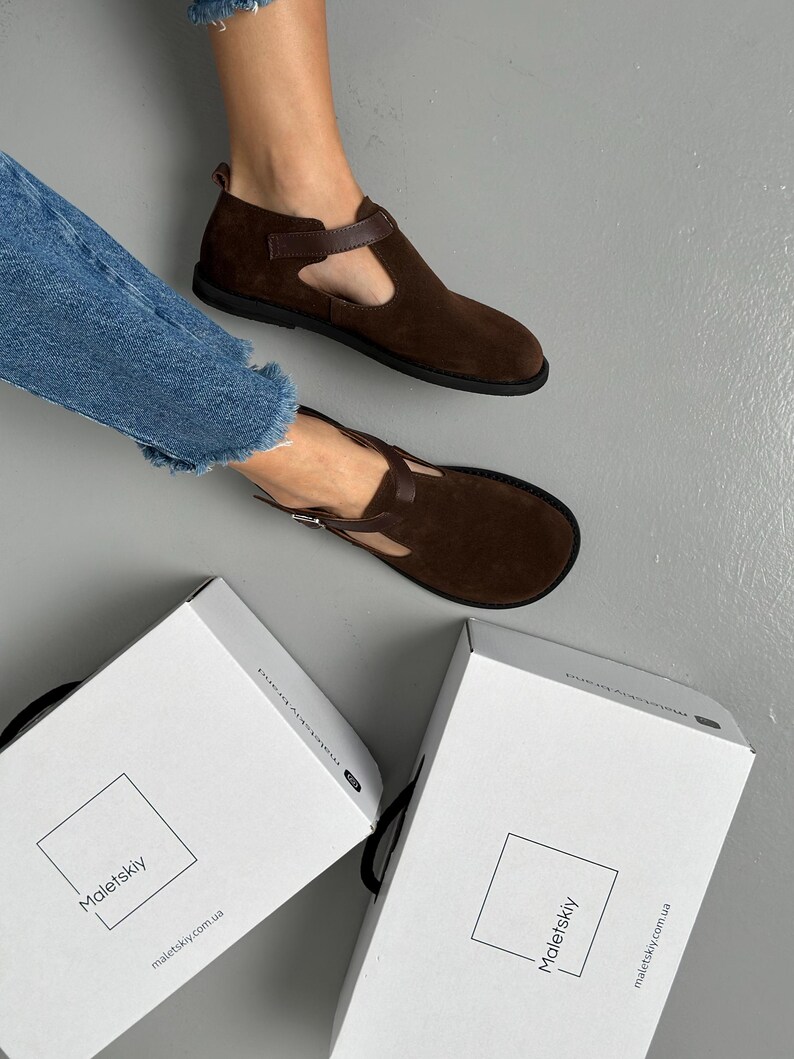 May include: Brown suede T-strap shoes with a dark brown strap and buckle closure. The shoes have a rounded toe and a black sole. Two white boxes with the brand name "Maletskiy" are visible.