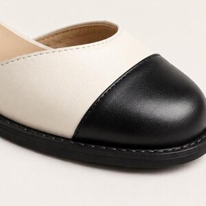 Puede incluir: Un zapato plano de dos tonos con una parte superior de color crema y una puntera redondeada negra. El zapato tiene costuras visibles a lo largo del borde de la suela y en la uni&oacute;n donde la puntera negra se encuentra con el cuerpo de color crema.