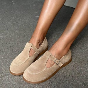 May include: A pair of tan suede Mary Jane style shoes with a buckle closure and a rounded toe. The shoes have a flat, brown sole. The image shows the shoes on a gray surface.