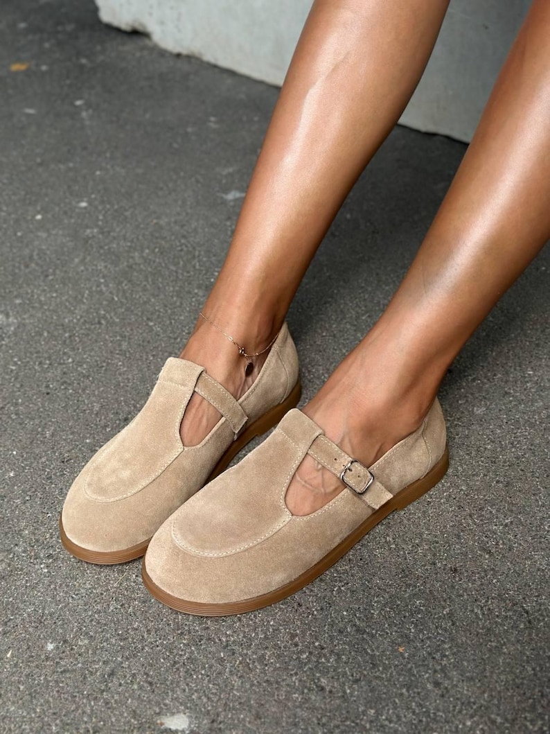 May include: A pair of tan suede Mary Jane style shoes with a buckle closure and a rounded toe. The shoes have a flat, brown sole. The image shows the shoes on a gray surface.
