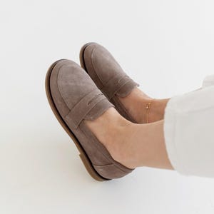 May include: A pair of taupe suede loafers with a classic penny loafer design. The shoes have a rounded toe and a brown sole. The image shows the shoes on a person's feet, with white pants in the background.