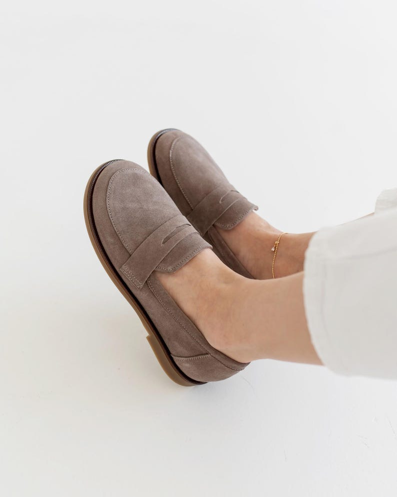 May include: A pair of taupe suede loafers with a classic penny loafer design. The shoes have a rounded toe and a brown sole. The image shows the shoes on a person's feet, with white pants in the background.