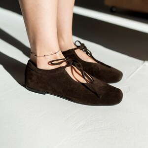 May include: A pair of dark brown suede flats with a lace-up design. The shoes have a rounded toe and a flat sole. A silver anklet with small beads is visible. The shoes are on a white surface.