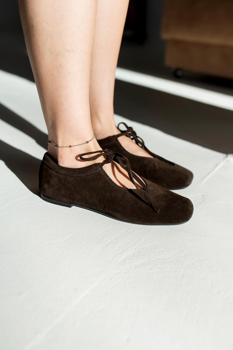 May include: A pair of dark brown suede flats with a lace-up design. The shoes have a rounded toe and a flat sole. A silver anklet with small beads is visible. The shoes are on a white surface.