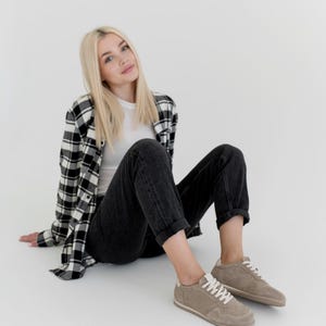Pu&ograve; includere: Una persona indossa una maglietta bianca, jeans neri e una camicia a quadri bianchi e neri. La persona indossa scarpe da ginnastica in pelle scamosciata marrone chiaro con lacci bianchi.