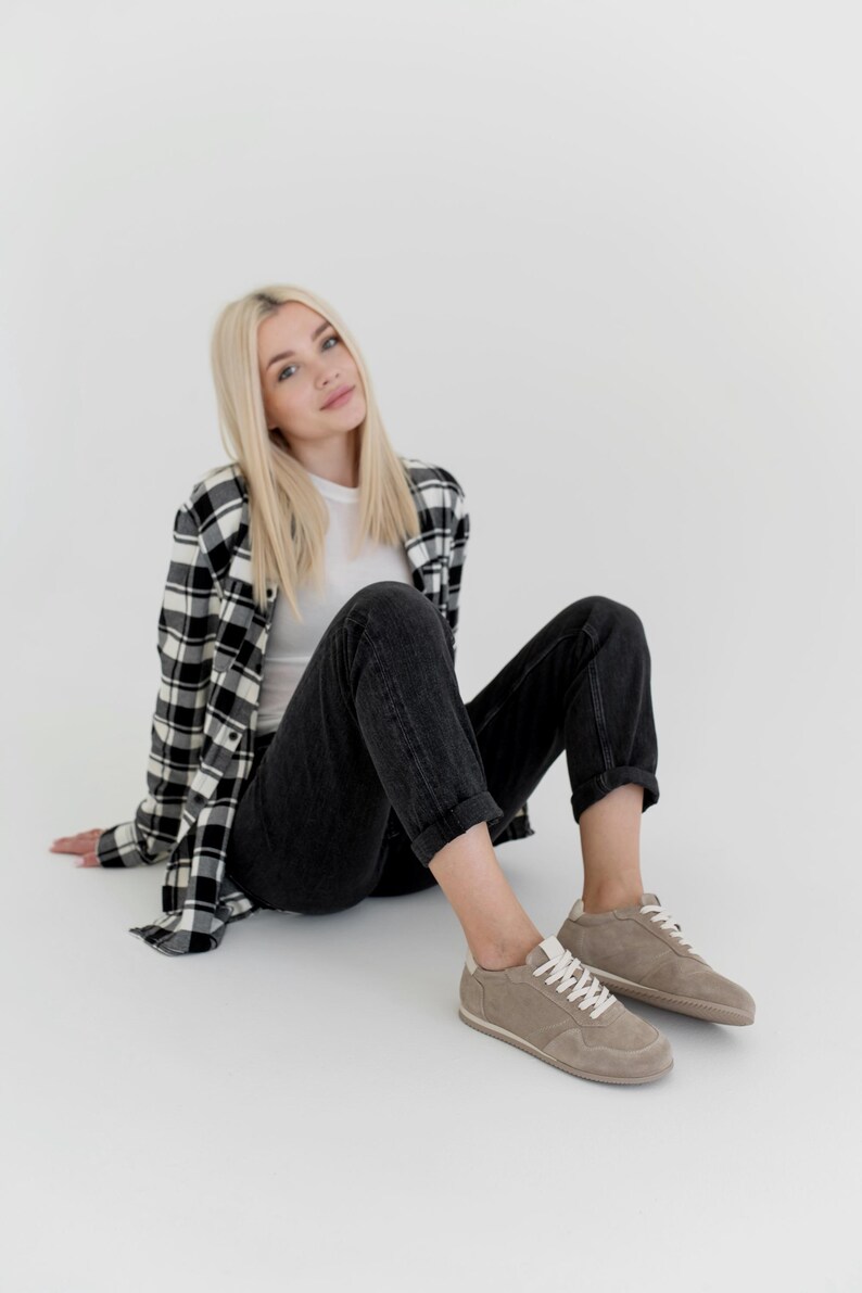 Pu&ograve; includere: Una persona indossa una maglietta bianca, jeans neri e una camicia a quadri bianchi e neri. La persona indossa scarpe da ginnastica in pelle scamosciata marrone chiaro con lacci bianchi.
