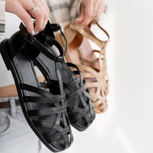 May include: Black leather sandals with multiple straps and a buckle closure are held up next to a pair of tan sandals. The black sandals have a flat sole, while the tan sandals have a higher heel. Both pairs of sandals are in focus.