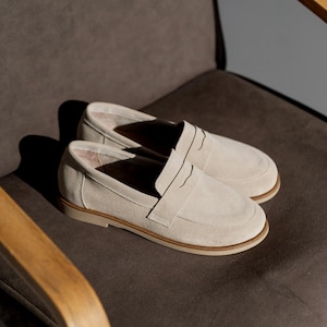 May include: A pair of light beige suede loafers with a slip-on design. The shoes have a rounded toe and a low, tan-colored sole. The loafers are displayed on a dark gray chair.