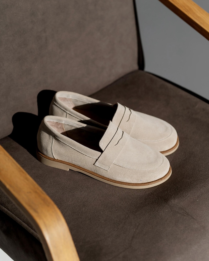 May include: A pair of light beige suede loafers with a slip-on design. The shoes have a rounded toe and a low, tan-colored sole. The loafers are displayed on a dark gray chair.