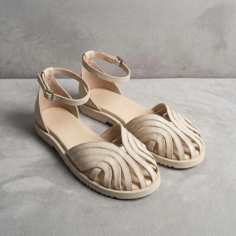 Puede incluir: Un par de sandalias de cuero beige con una correa en el tobillo y cierre de hebilla. Las sandalias tienen una suela plana y un dise&ntilde;o decorativo de correas curvas en la parte superior. Los zapatos se muestran sobre una superficie gris.