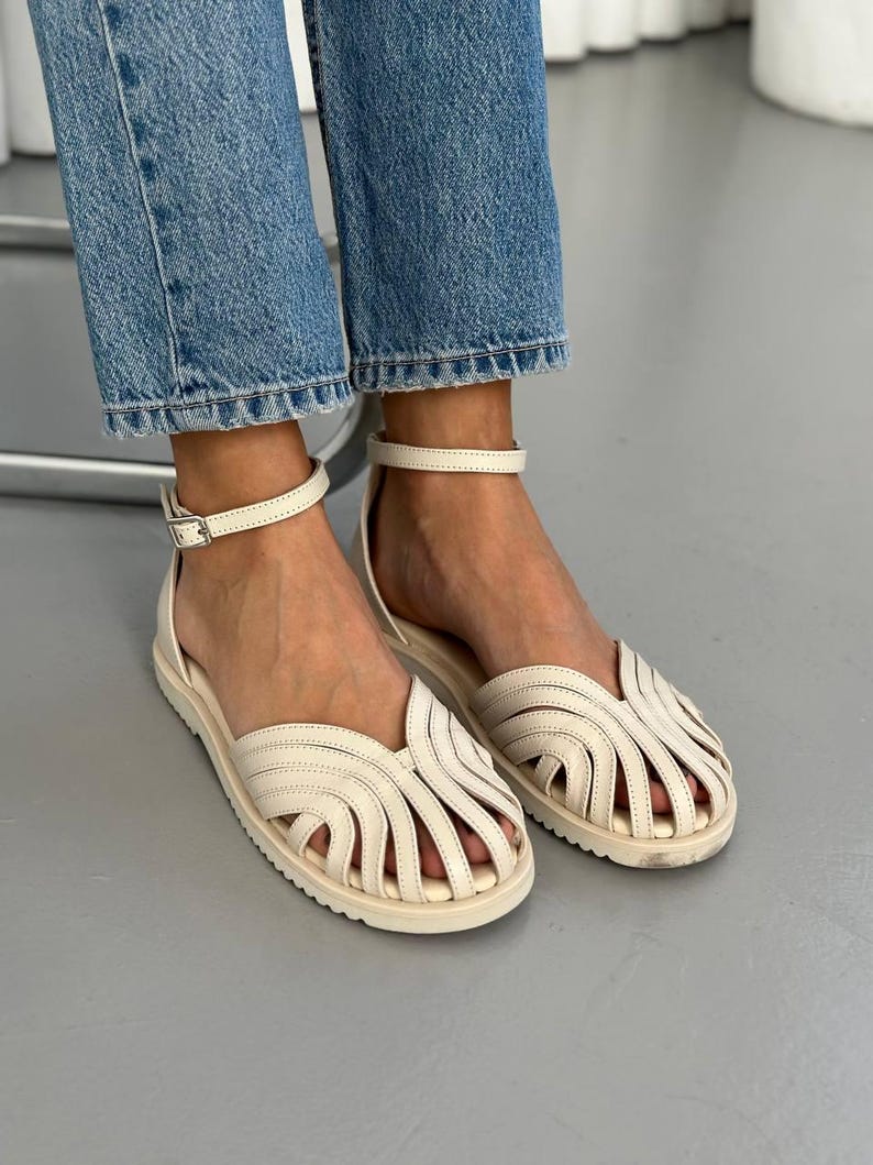 Puede incluir: Sandalias color crema con dise&ntilde;o tejido y correas en el tobillo. Las sandalias tienen suela plana y cierre de hebilla. La persona lleva vaqueros azules.