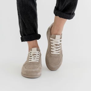Pu&ograve; includere: Un paio di sneakers in camoscio beige con lacci bianchi e linguetta bianca. Le scarpe sono mostrate da una prospettiva bassa, con i jeans grigio scuro del portatore risvoltati alle caviglie. Lo sfondo &egrave; bianco.