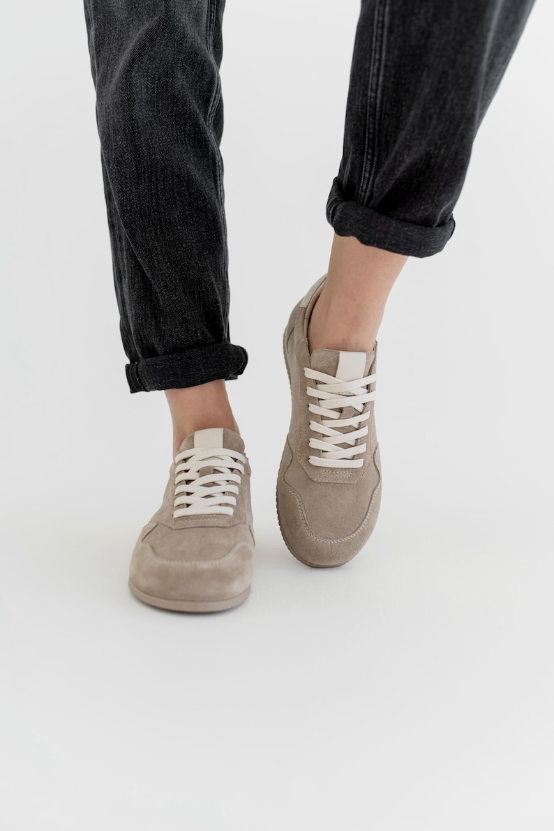 Pu&ograve; includere: Un paio di sneakers in camoscio beige con lacci bianchi e linguetta bianca. Le scarpe sono mostrate da una prospettiva bassa, con i jeans grigio scuro del portatore risvoltati alle caviglie. Lo sfondo &egrave; bianco.