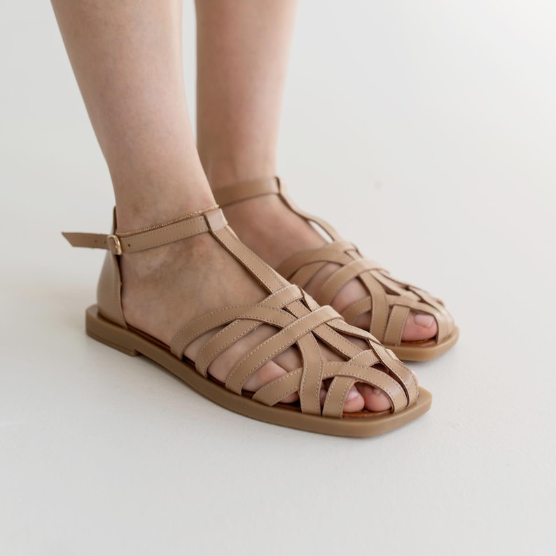 May include: Tan leather sandals with a woven design across the foot and a T-strap. The sandals have a flat sole and an adjustable ankle strap with a buckle. The footwear is displayed against a white background.