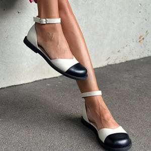 May include: A pair of two-tone flat shoes featuring a cream-colored body and a rounded black toe cap. The shoes have a low-cut side profile, a thin ankle strap with a silver-toned metal buckle, and a flat black sole for a simple, functional design.