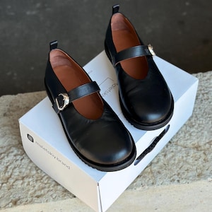 Può includere: Un paio di scarpe Mary Jane in pelle nera con cinturino e fibbia. Le scarpe sono su una scatola bianca con la scritta "maletskiy.brand". Hanno la punta arrotondata e la suola spessa. Stile classico.