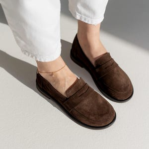 May include: A pair of dark brown suede loafers featuring a classic penny slot design across the vamp, a rounded toe, and a flat black sole. The shoes are worn with white trousers and a delicate gold chain anklet with a small charm.