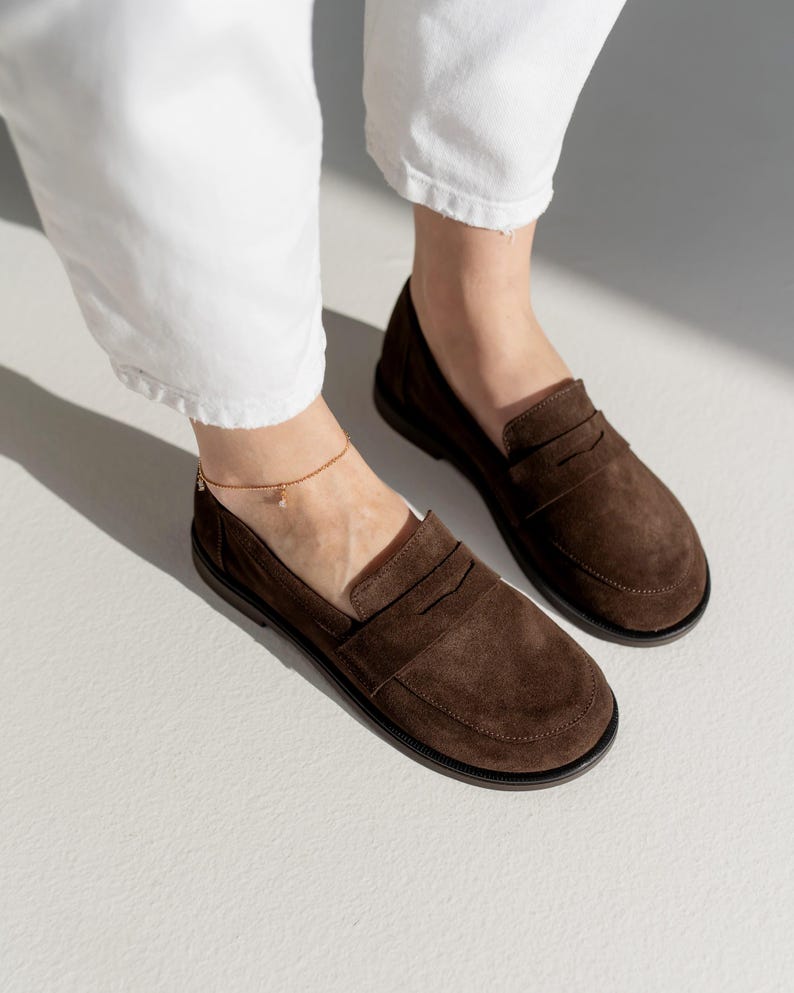 May include: A pair of dark brown suede loafers featuring a classic penny slot design across the vamp, a rounded toe, and a flat black sole. The shoes are worn with white trousers and a delicate gold chain anklet with a small charm.