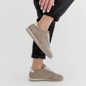 Pu&ograve; includere: Un paio di sneakers in camoscio color talpa con lacci bianchi e un rinforzo bianco sul tallone. Le sneakers hanno un profilo basso e una palette di colori neutri, adatte all'abbigliamento casual. Le suole sono beige chiaro.