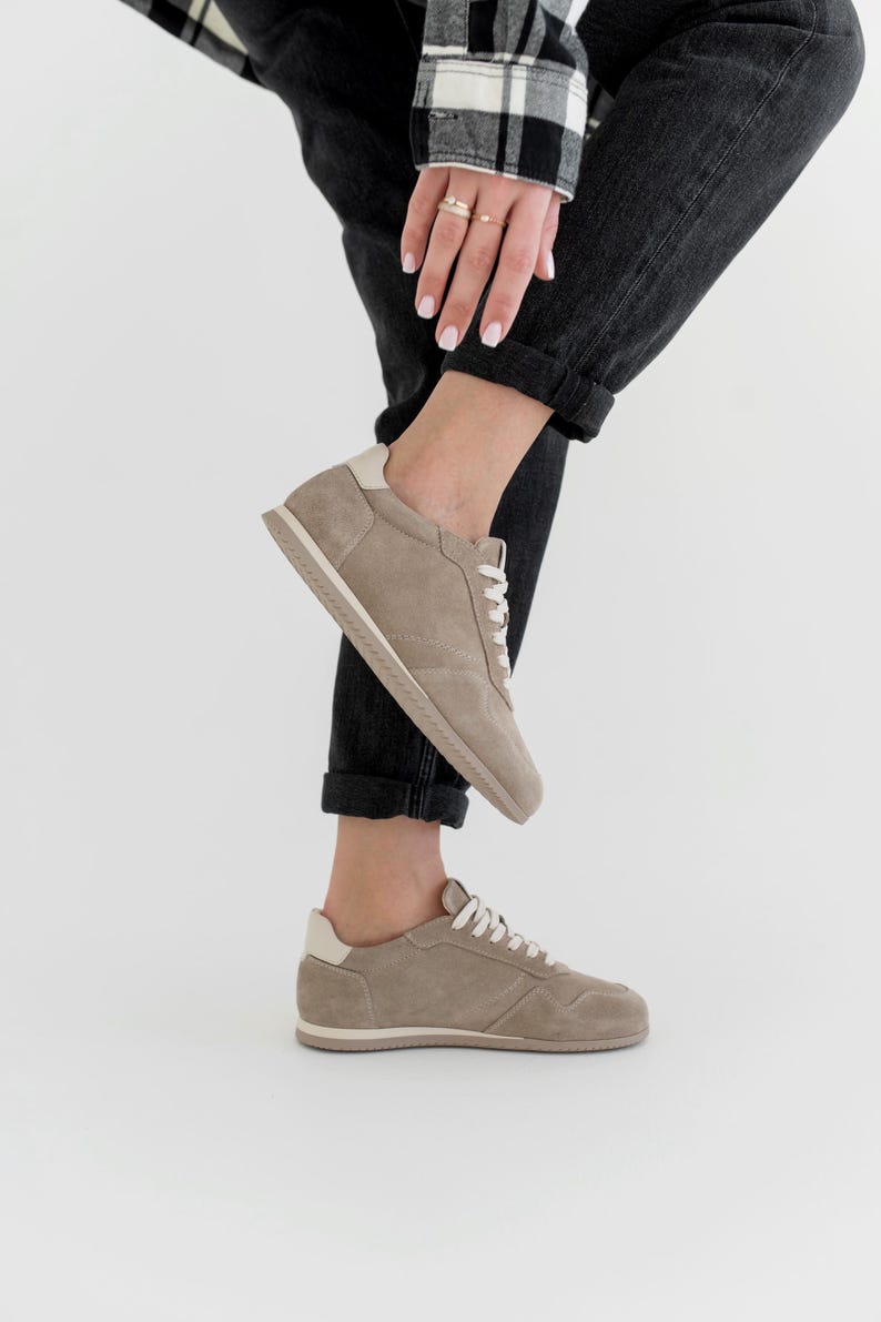 Pu&ograve; includere: Un paio di sneakers in camoscio color talpa con lacci bianchi e un rinforzo bianco sul tallone. Le sneakers hanno un profilo basso e una palette di colori neutri, adatte all'abbigliamento casual. Le suole sono beige chiaro.