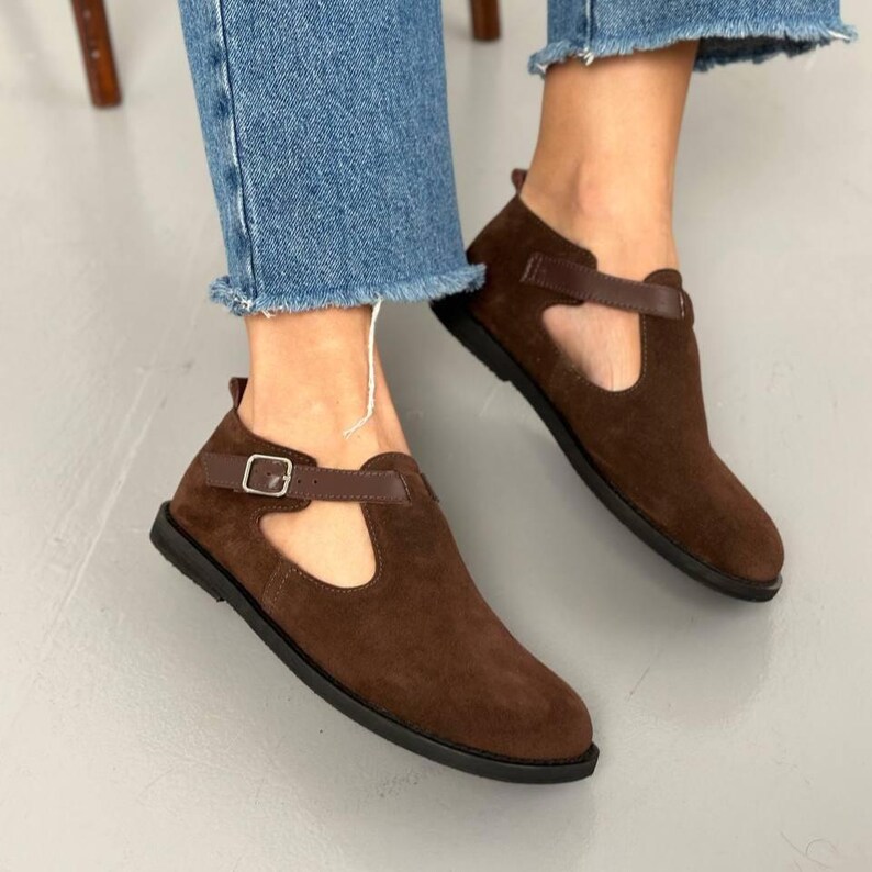 May include: Brown suede ankle boots with a strap and buckle closure. The shoes have a cutout design and a dark brown sole. The boots are worn with blue jeans.