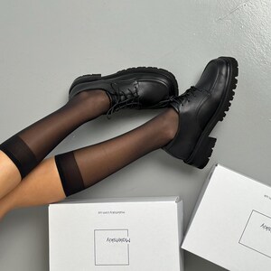 May include: Black leather lace-up shoes with a chunky sole. The shoes are paired with sheer black knee-high socks. Two white boxes with the text "Maletskiy" are in the background.