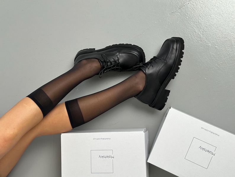May include: Black leather lace-up shoes with a chunky sole. The shoes are paired with sheer black knee-high socks. Two white boxes with the text "Maletskiy" are in the background.