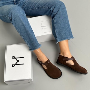 May include: Brown suede T-strap flats with a buckle closure. The shoes are paired with a white box with the brand name "Maletskiy" and a logo. The model is wearing blue jeans.