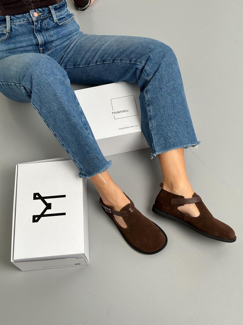 May include: Brown suede T-strap flats with a buckle closure. The shoes are paired with a white box with the brand name "Maletskiy" and a logo. The model is wearing blue jeans.