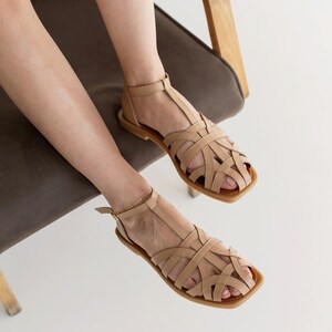 May include: Tan leather sandals with a woven design. The sandals have a square toe and a strap that goes around the ankle. The sandals are sitting on a brown chair.